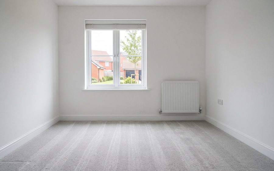 An empty carpeted room with bare walls, clean skirtings and a clear window